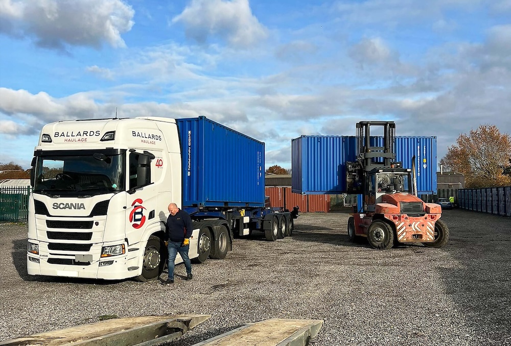Shows Ballards expanding self storage facilities to meet growing demand - a truck carrying a container and a forklift carrying another one on a gravelled area